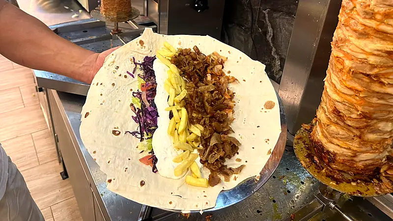 Rozložený dürüm s masem, hranolkami, zelím a omáčkou vedle grilu s kebabem