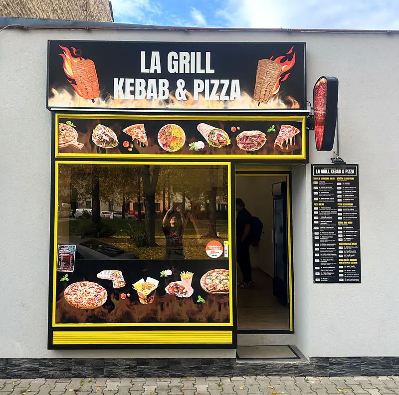 Provozovna La Grill z&nbsp;chodníku: černý štít s&nbsp;nápisem, výloha ve žlutém rámu, ceník na zdi a&nbsp;červený světelný kebab